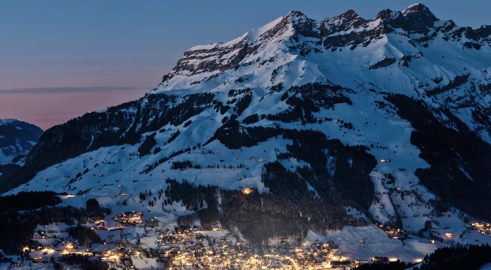 Twilight view of a glowing alpine village nestled beneath dramatic snow-covered peaks, highlighting why SWISStours is the Best place to buy swiss travel pass for winter travel.
