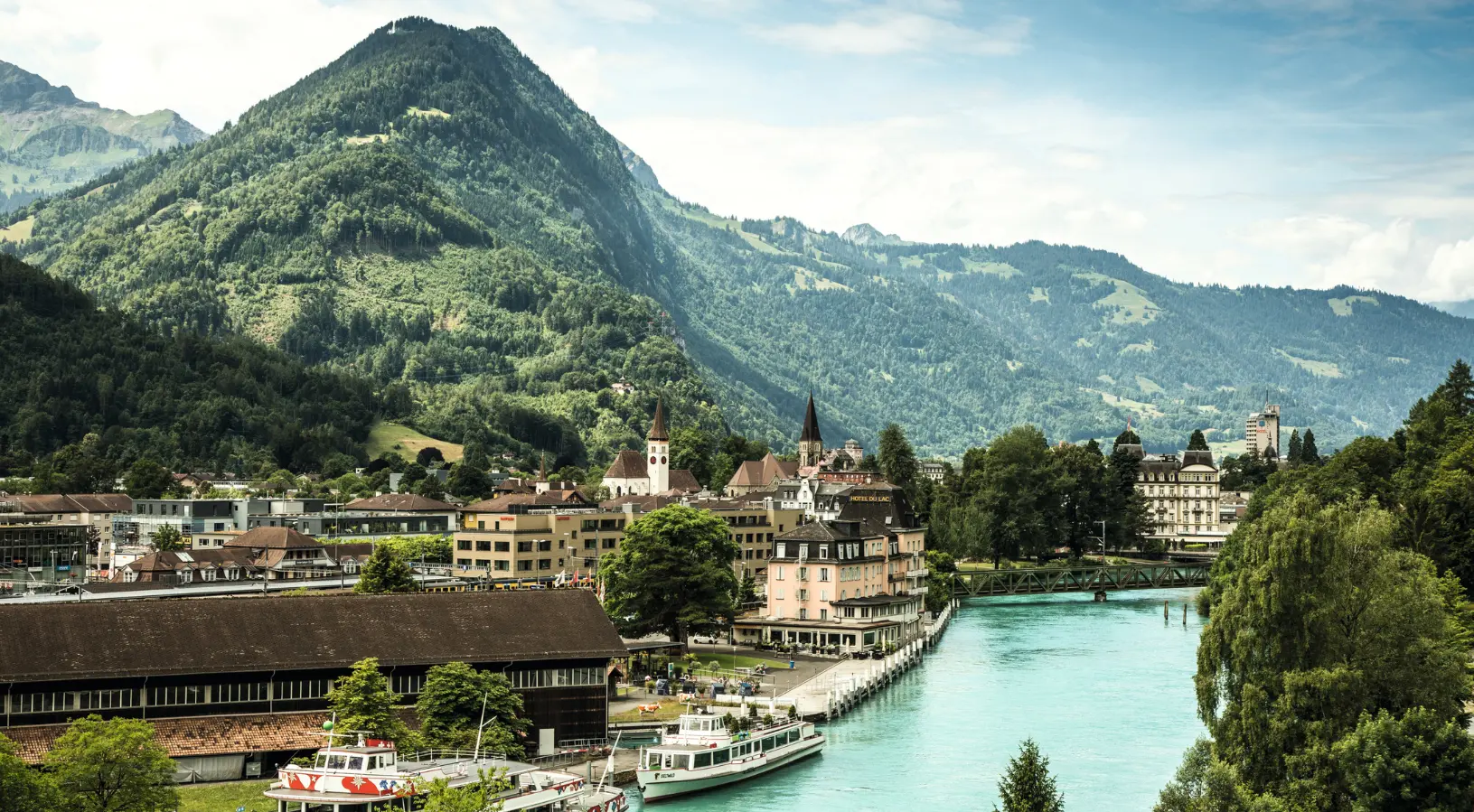 Scenic view of Interlaken with the turquoise Aare River, historic buildings, and surrounding Swiss Alps, a picturesque highlight of a romantic Switzerland Tour Package for couple.