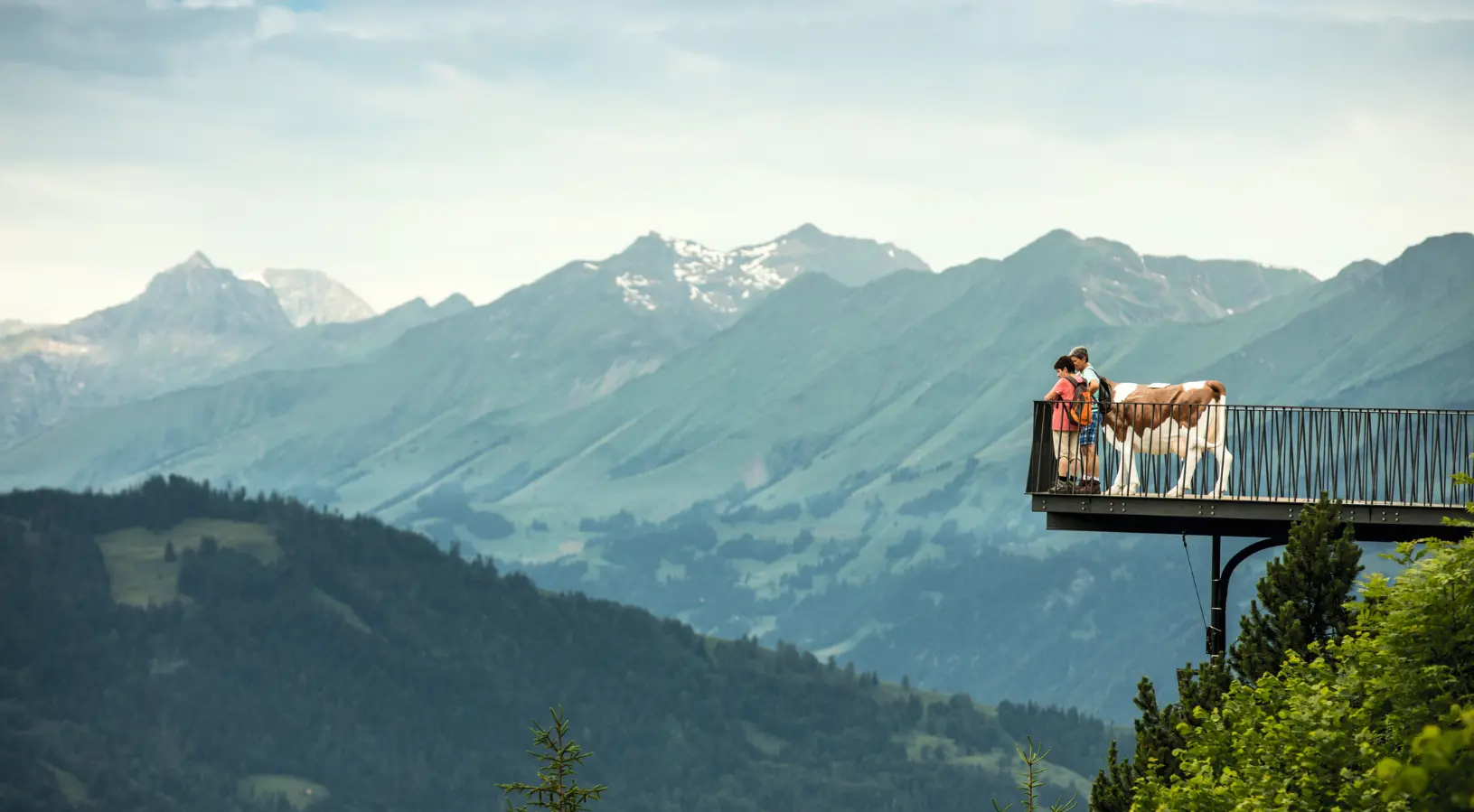 A family standing on the metal viewing platform at Grindelwald-First next to a cow statue, a popular adventure day trip among things to do in Interlaken.