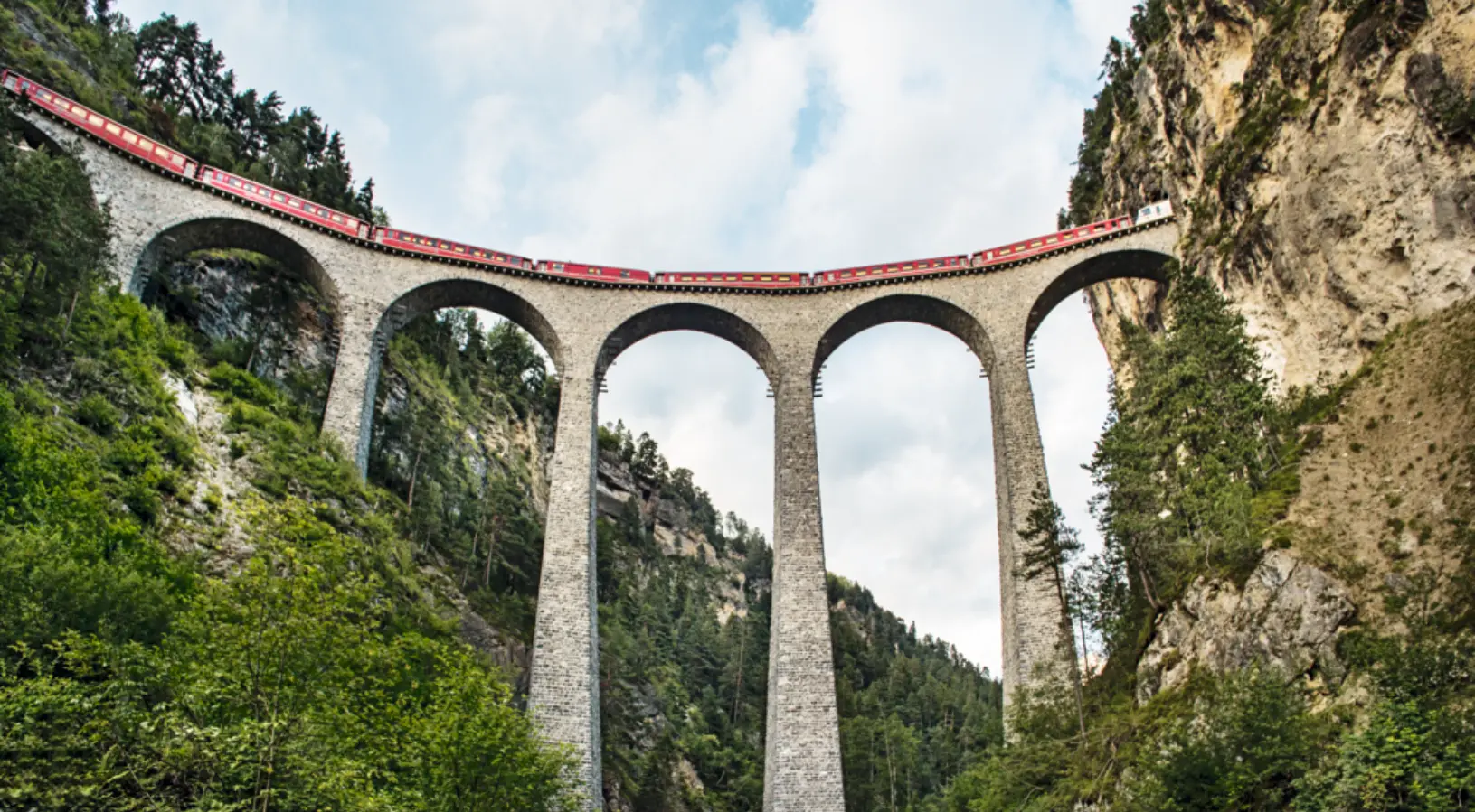 The iconic red Bernina Express train crosses the majestic Landwasser Viaduct, a stunning stone bridge curving through the Swiss Alps between cliffs and lush green forests. This breathtaking scene showcases one of Switzerland’s most famous rail journeys, making it a highlight in the Swiss Travel Pass Guide for exploring the country’s world-renowned scenic routes.