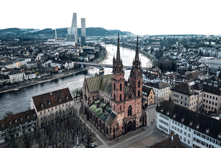 Aerial view of Basel Minster in Basel, Switzerland, showcasing its striking Gothic architecture, twin towers, and vibrant red sandstone façade against the backdrop of the city's charming rooftops and the Rhine River.