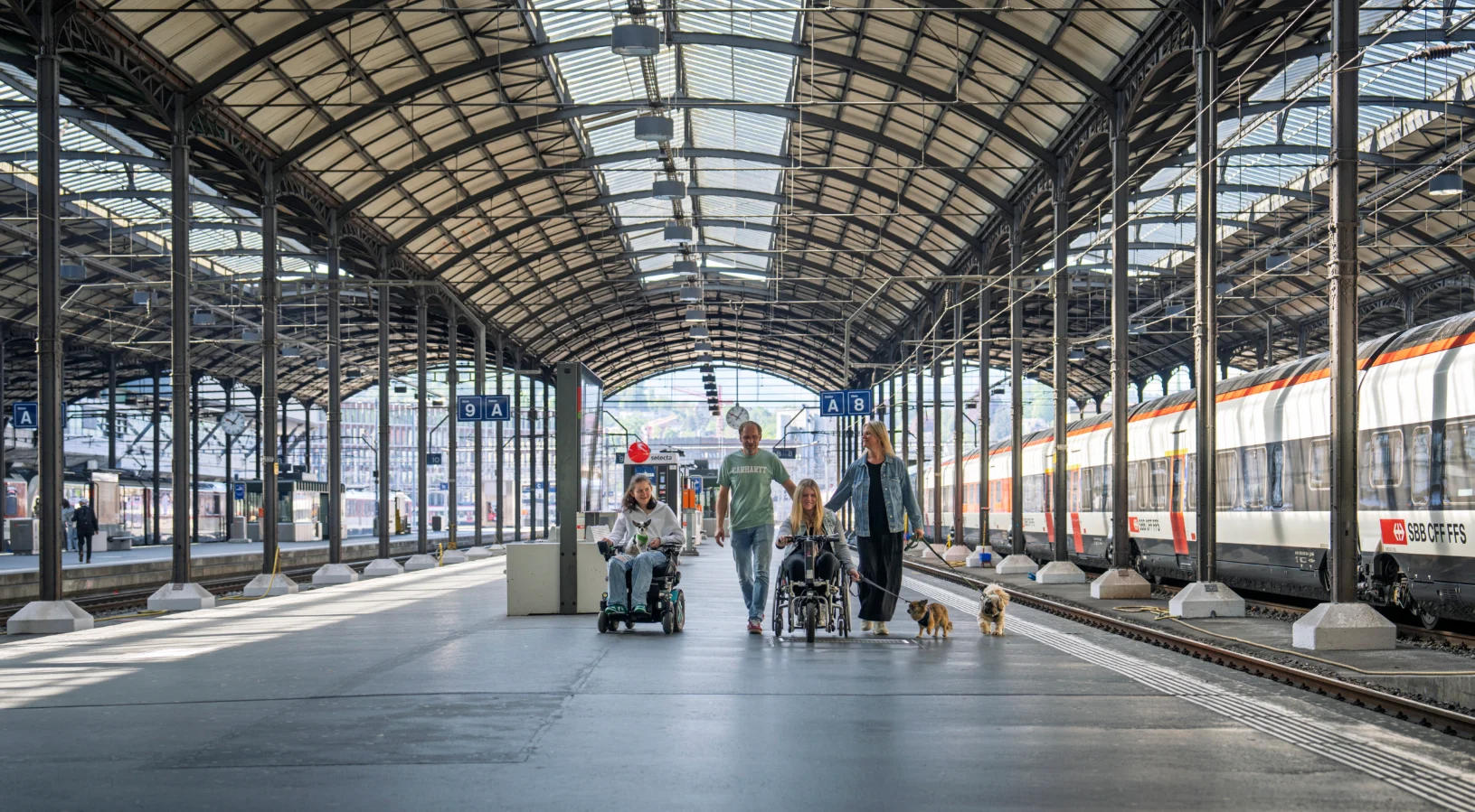 Travellers walking along a platform under the arched roof of Lucerne railway station beside a Swiss train, illustrating how to buy the Swiss Travel Pass from India