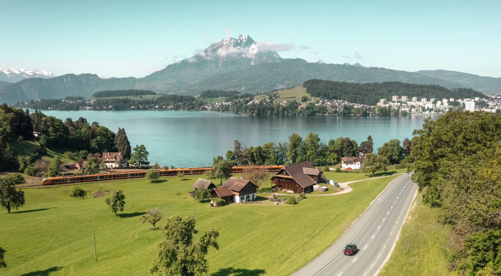 A Swiss train running through green meadows beside Lake Lucerne with Mount Pilatus behind, illustrating what decides the price of a Swiss Travel Pass